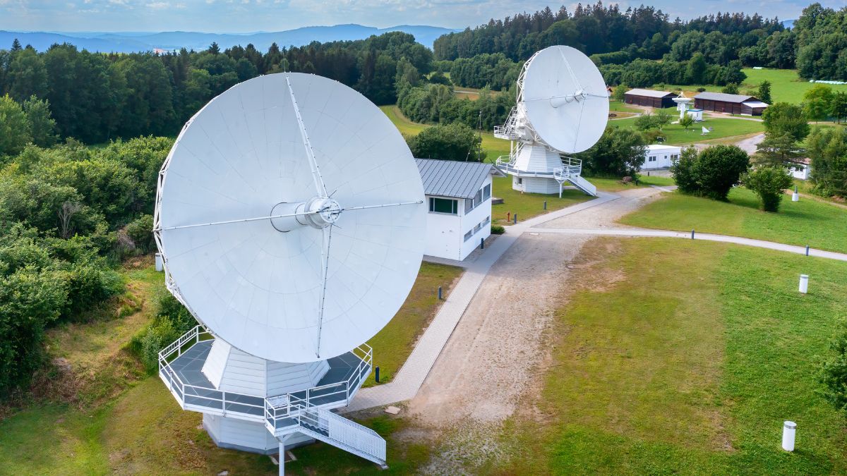 Zwei Radioteleskope. Dazwischen ein Kontrollgebäude mit Giebeldach. Im Hintergrund eine typische Mittelgebirgslandschaft. (verweist auf: Meilensteine in der Geophysik 4/4)
